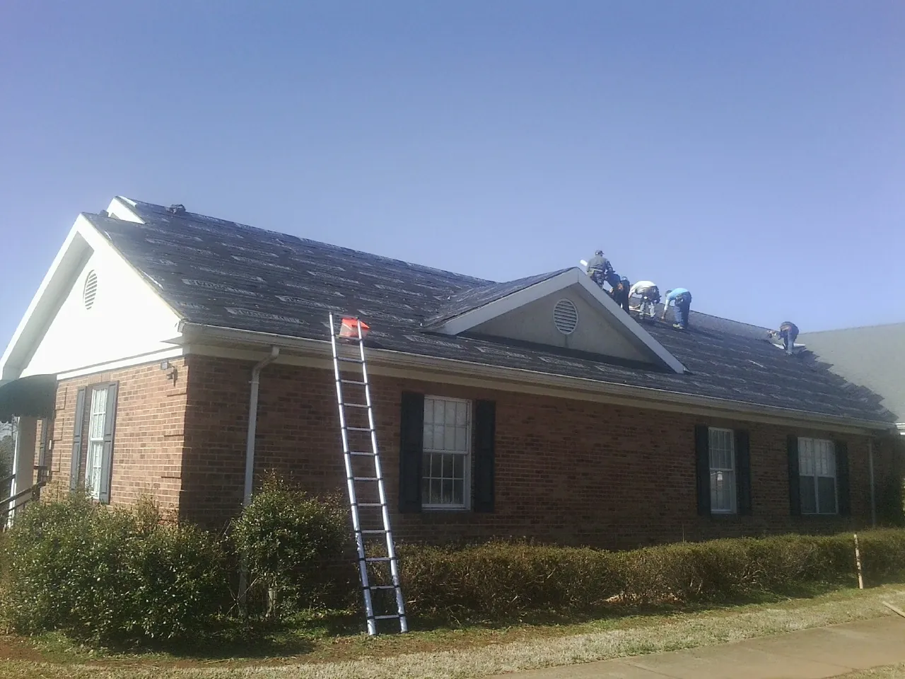 Crew tearing shingles off a brick ranch roof with a ladder set — Emergency Roof Repair work by our crew in Rembert
