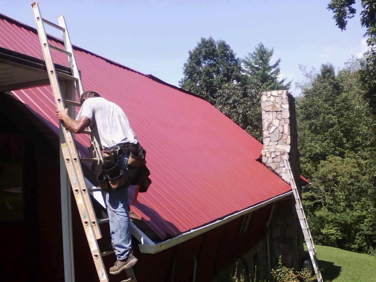 Roofer on a ladder beside a red metal roof and stone chimney — Soffit & Fascia Repair by Roofing Brothers in Rembert